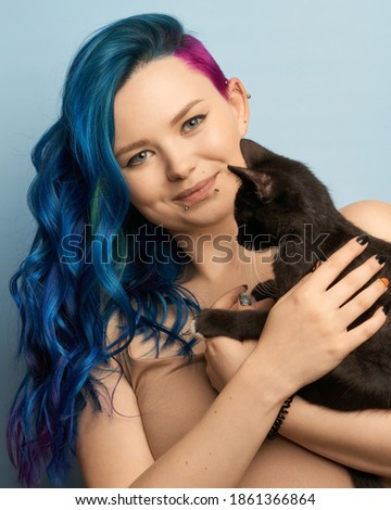 Girl with creative blue coloring and a rainbow in her hair on a blue background holds black cat. Modern minimalistic bright photography for advertising and social networks