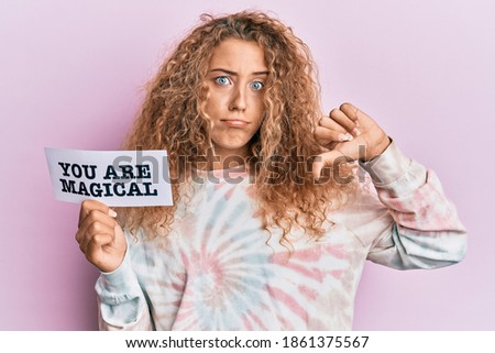 Beautiful caucasian teenager girl holding you are magical banner with angry face, negative sign showing dislike with thumbs down, rejection concept 