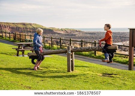 A young brother and sister playing on a seesaw in a park at Crail, Fife, Scotland. This park is on the coastal trail and is in a very beautiful location. Royalty-Free Stock Photo #1861378507