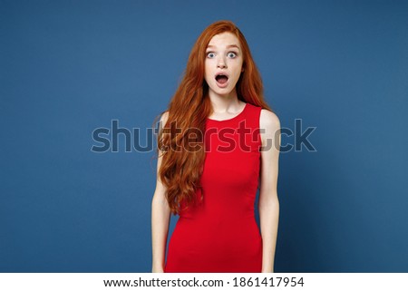 Shocked amazed worried young redhead woman 20s wearing bright red elegant evening dress standing keeping mouth open looking camera isolated on blue color wall background studio portrait