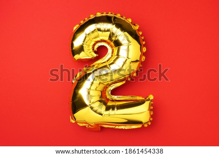 Creative layout. Golden foil balloon number and digit two 2. Birthday greeting card. Anniversary concept. Top view. Copy space. Stylish gold numeral over red background. Numerical digit