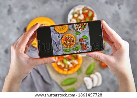 Taking photo of food with hands holding mobile phone with picture of vegan baked Red kuri squash vegetable filled with bell pepper, tomatoes and mushrooms surrounded by ingredients