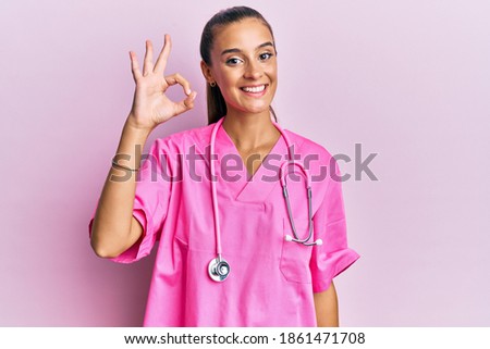 Young hispanic woman wearing doctor uniform and stethoscope smiling positive doing ok sign with hand and fingers. successful expression. 