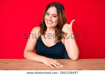 Young beautiful caucasian woman wearing casual clothes sitting on the table smiling happy and positive, thumb up doing excellent and approval sign 