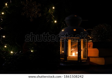 White lantern with a burning candle. Books, Christmas tree branch and lights on background. Night dark picture. Christmas mystery mood.