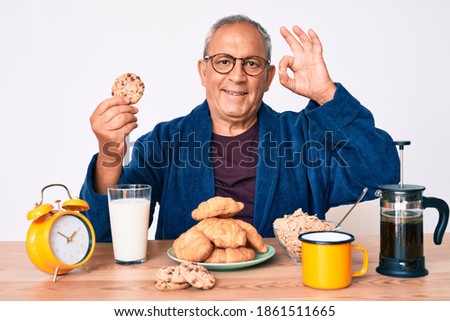 Senior handsome man with gray hair sitting on the table eating breakfast in the morning doing ok sign with fingers, smiling friendly gesturing excellent symbol 