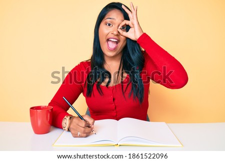 Beautiful latin young woman with long hair writing a book sitting on the table smiling happy doing ok sign with hand on eye looking through fingers 
