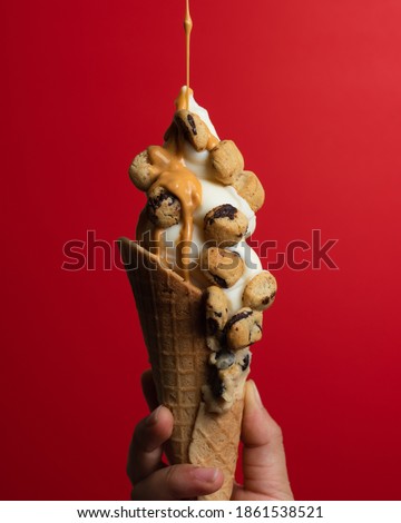 A hand holding an ice cream cone topped with cookie dough bites while caramel sauce is streaming down from the top of the frame