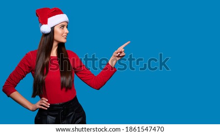 Excited young woman in Christmas Santa hat showing or pointing with finger away on blue isolated background