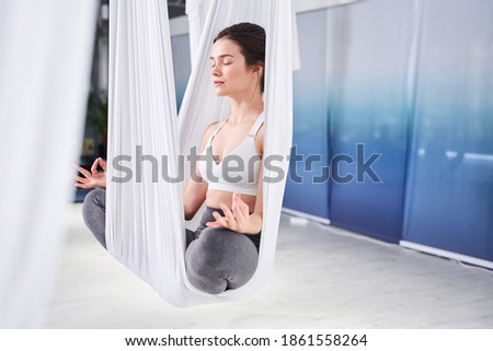 Photo of the young mindful caucasian woman sitting at the lotus pose at the hammock and meditating at the light yoga studio. Relax, breathe easy, seat pose, gym, healthy lifestyle concept