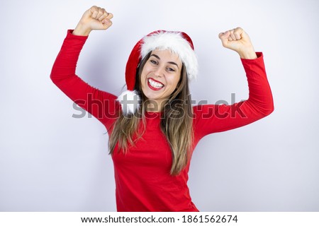 Young beautiful woman wearing a Santa hat over white background very happy and excited making winner gesture with raised arms, smiling and screaming for success.