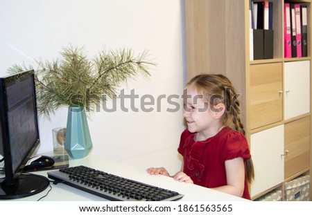 A little girl in red and white clothes looks at the monitor and smiles. The girl is talking on a video call on the computer. Distant life.
