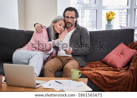 Coronavirus has affected our financial capabilities. Senior sad woman lies on her husband's shoulder while sitting at the sofa and looking with sadness at the bills or payments. Poorness concept