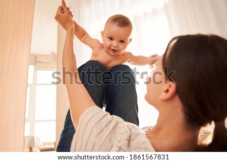 My active boy! Young attractive mother laying at the back and playing with her newborn at the bedroom while spending great time together. Stock photo
