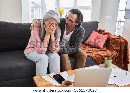 Senior concerned man wearing glasses consoles his upset crying wife and embracing her at home at the sofa after viewing utility bills. Poorness concept. Stock photo