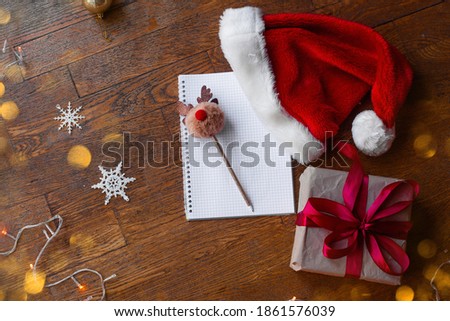 Flatlay on wooden brown floor New Year's mood. Writing Christmas wishes to Santa Claus. On textured background lies notepad in cage, deer pompom pen, gift with red ribbon, craft paper, hat. Copy space