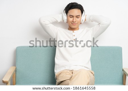 Asian businessman wearing wireless headphones to listen to music while sitting on sofa at home
