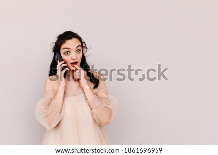 Beautiful woman of European appearance talking in surprise on the phone against a beige background. High quality photo