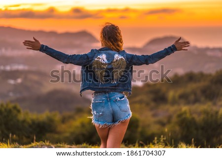 Lifestyle, a Caucasian girl with a denim jacket and an angel wings print on the back, with denim shorts. With open arms atop a mountain in a gorgeous summer sunset