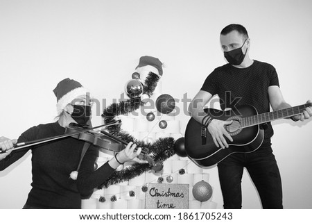 man and woman playing musical instruments near christmas tree made of toilet paper rolls. holidays at home quarantine in 2020. lockdown. girl and boy wearing face masks to protect from coronavirus. 