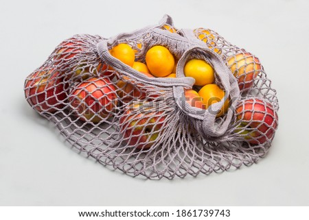 Hand holds string bag mesh with red apples and tangerines. Ecological concept. Gray background. Top view
