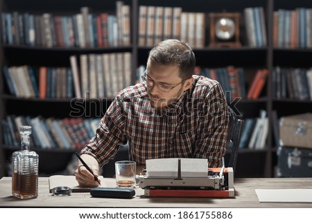 A young writer with glasses sitting at a Desk writes a story with a fountain pen next to a typewriter and alcohol against the background of a library Cabinet Royalty-Free Stock Photo #1861755886