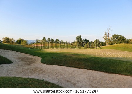 Golf terrain with short trimmed grass and sand bar with yellow flag gently swaying in the wind, surrounded by green woods                 