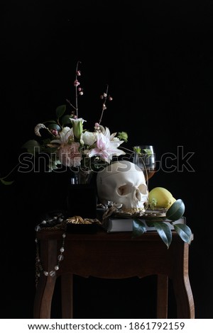 Close up still life picture inspired by 17th century Dutch painting. There are flowers, fruits, jewelry, a glass of wine and a skull on wooden table, in front of a black background.