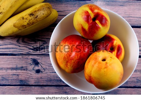 A top view closeup of juicy nectarines in a white bowl next to bananas on a wooden table