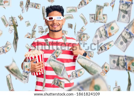 Handsome latin american young man wearing 3d glasses and eating popcorn at the movies pointing finger to one self smiling happy and proud