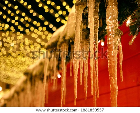 Icicles hang from the roof of a red house decorated with Christmas tree decorations. Selective focus, blurred background, bokeh of golden lights.