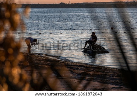 In the distance, a photographer shoots a young couple on the shore of a sandy beach, calm sea at sunset in the resort town of Gelendzhik. The view through the tall bushes. Russia, Black sea coast