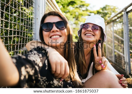 Two cheerful girls taking off a selfie while sitting together on a bridge and showing a gesture of peace outdoors - Two charming smiling woman taking selfie on smartphone.