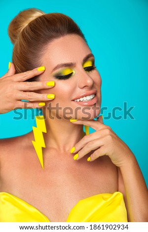 Beautiful girl with bright makeup and yellow manicured nails isolated blue background. Closeup portrait of blonde with lightning flat earrings jewelry. 