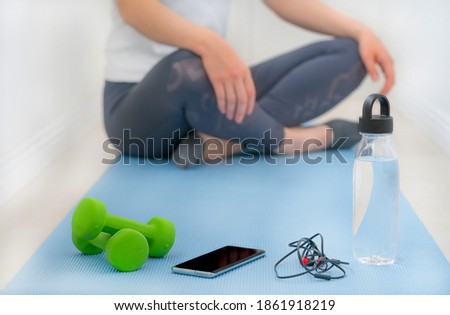 Sporty woman doing workout or yoga at home stock photo. Close-up on sport mat, dumbbells, bottle with water, headphones and smartphone