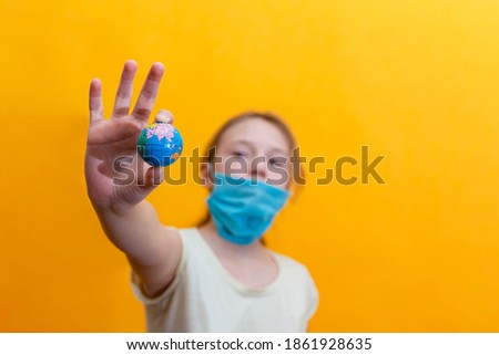 A girl with a full-face mask holds a toy earth in her right hand on an orange background.