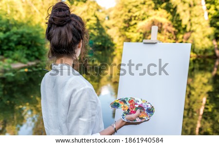 girl artist summer park pond, background forest water shore trees, draws picture, view from front, hand palette with paints easel. Clothes white shirt. White blank canvas looking at landscape.