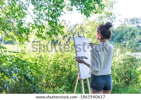 woman artist summer forest, background bushes trees, begins paint picture white canvas, in hand brush color palette of paints, easel. Hair in a bun. Free space rear view
