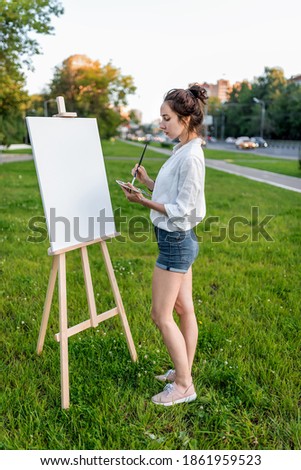 woman artist city summer, background car road, green lawn, begins paint picture on white canvas, hand brush color palette of paints, easel. Clothes white shirt shorts. Hair bun