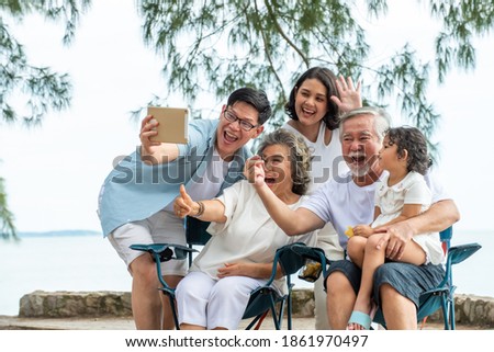 Group of Multi generation Asian family using digital tablet video call with their family friend on the beach in summer day. Happy big family enjoy and having fun together in summer holiday vacation