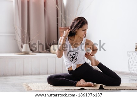 Flexible concentrated beautiful young brunette doing eka pada rajdakapotasana sitting on a rug on the floor in sportswear in an empty room. Concept of improving joints and rejuvenating the body