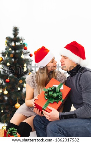 A loving couple looks at each other in Christmas hats with Christmas gifts, against the background of a Christmas tree