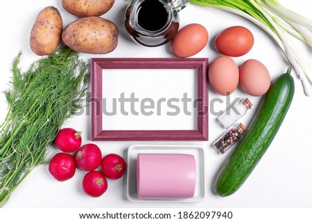 Ingredients to make Okroshka cold soup. Sausage, radish, potato, cucumber, eggs, kvass, herbs and spices around the picture frame on the white table. Top view