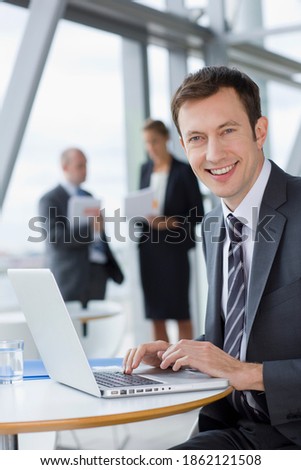 A vertical portrait of a smiling businessman in formal suit sitting at a table in the office lobby while using a laptop