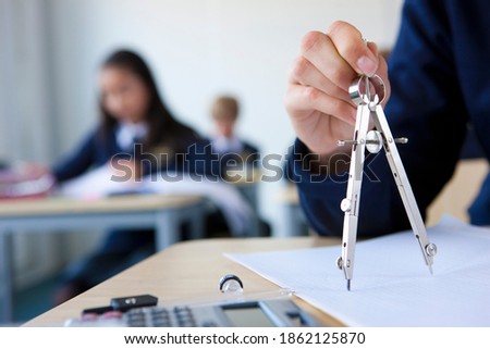 A close up shot of a student's hand using a compass on a desk in a classroom.