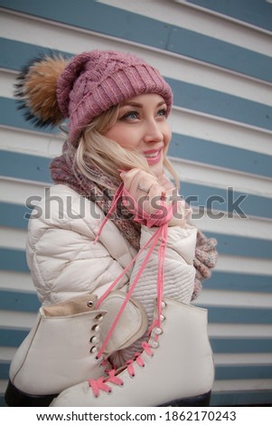 Pretty blond woman in pink hat, sweater, hat and white jacket posing with skates against the white and blue wall, wjman walking to the rink 