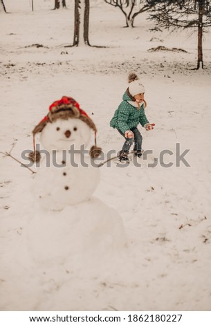 Little girl in a green and dot jacket and white hat in a snowy winter park with a snowman. Games, snow, fun, nature, smile, holiday. Children play in snow. Outdoor fun on cold winter day.