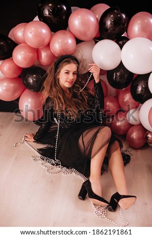 A party girl with long curly hair, in a black shiny dress in the image of the doll, sits among the balloons.