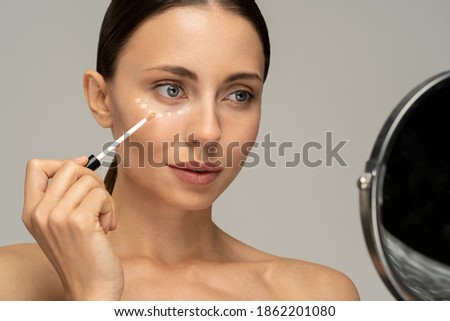 Woman with natural makeup applying corrector on flawless fresh skin, doing make up, looking at mirror. Girl after shower put concealer under eye area. Beauty face, skin care. Copy space, advertising. Royalty-Free Stock Photo #1862201080