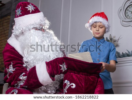 Santa Claus and a boy in a red hat are sitting by the Christmas tree, the child looks into Santa's bag and takes out gifts, Christmas and New Year, winter holidays.